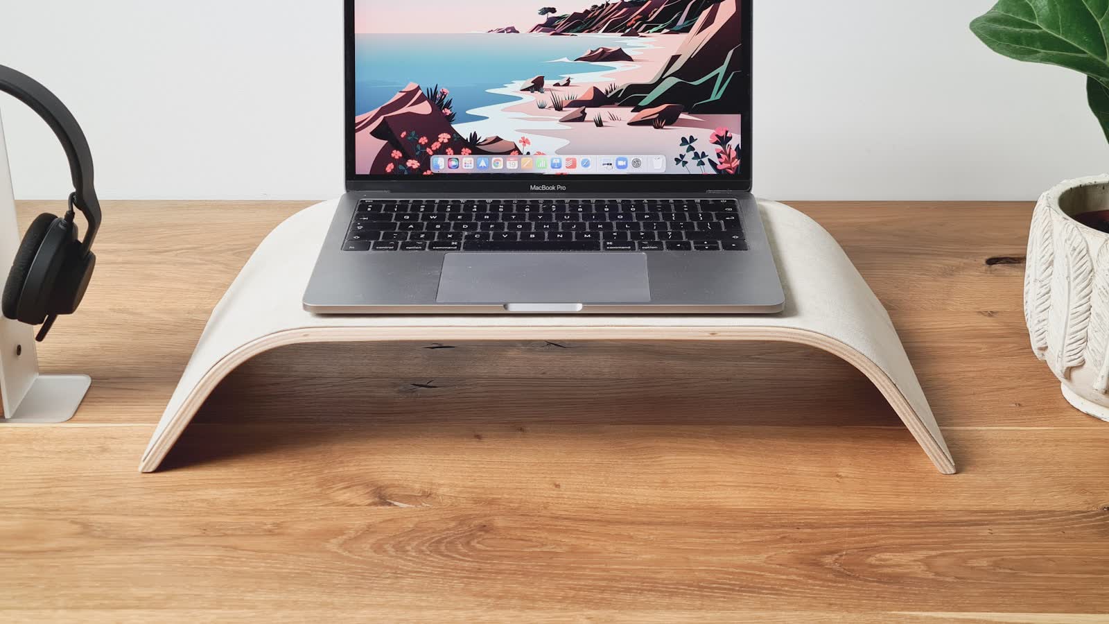 A laptop raised on a curved stand beside a potted plant